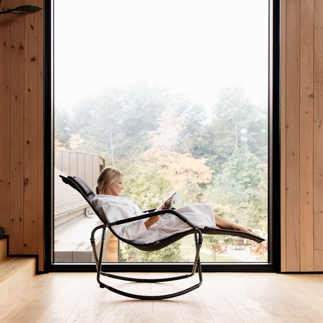 Femme détendue en peignoir, lisant dans un fauteuil inclinable moderne, avec une vue sur un paysage automnal à travers une grande baie vitrée dans un intérieur en bois.